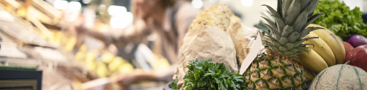 Closeup of shopping cart at supermarket full of food, fruit and vegetables while in background woman taking product off the shelves.